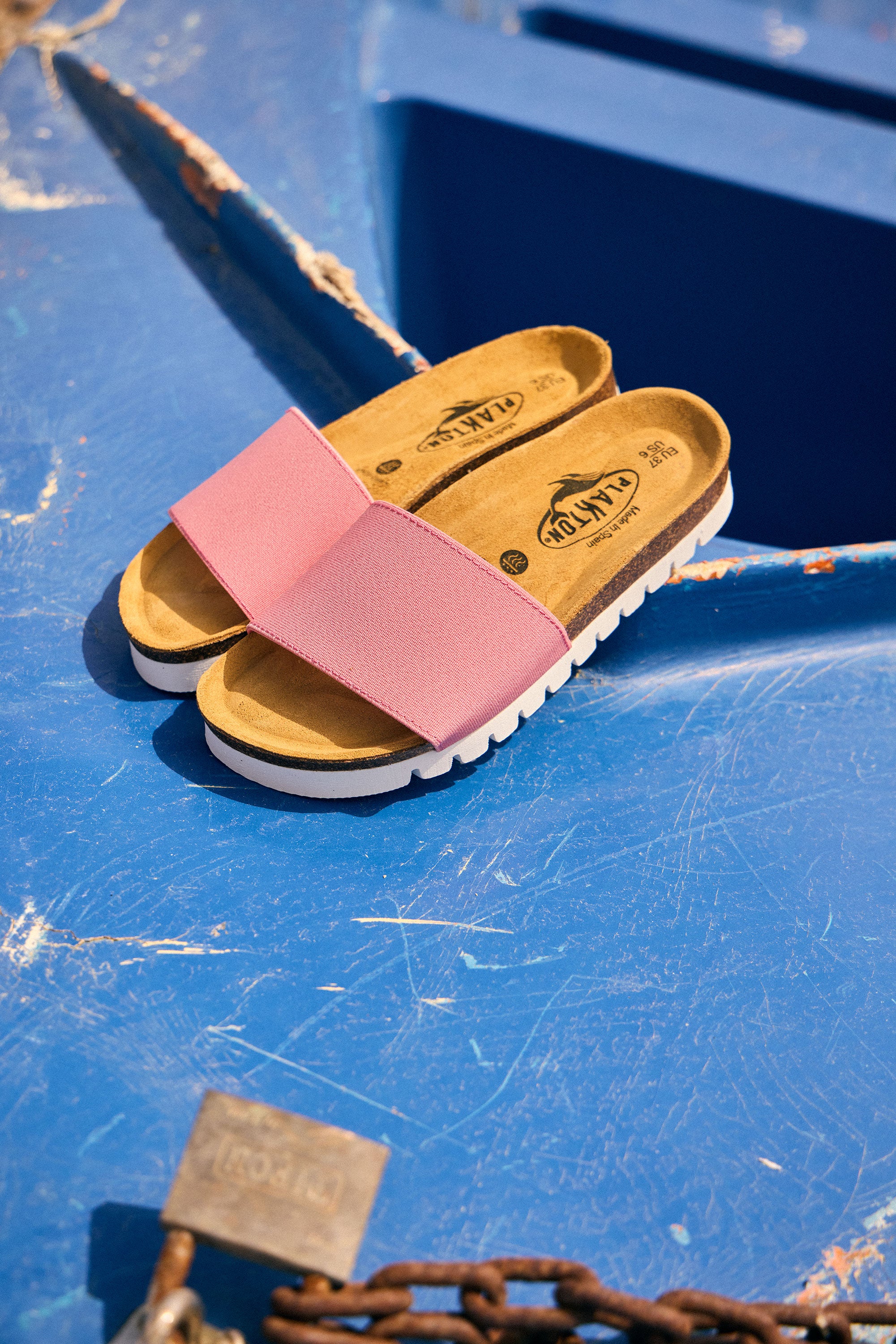 Plakton Barcadi pink flat sandals with elastic strap and cork footbed placed on a bright blue boat, highlighting comfortable handmade summer sandals from Spain.