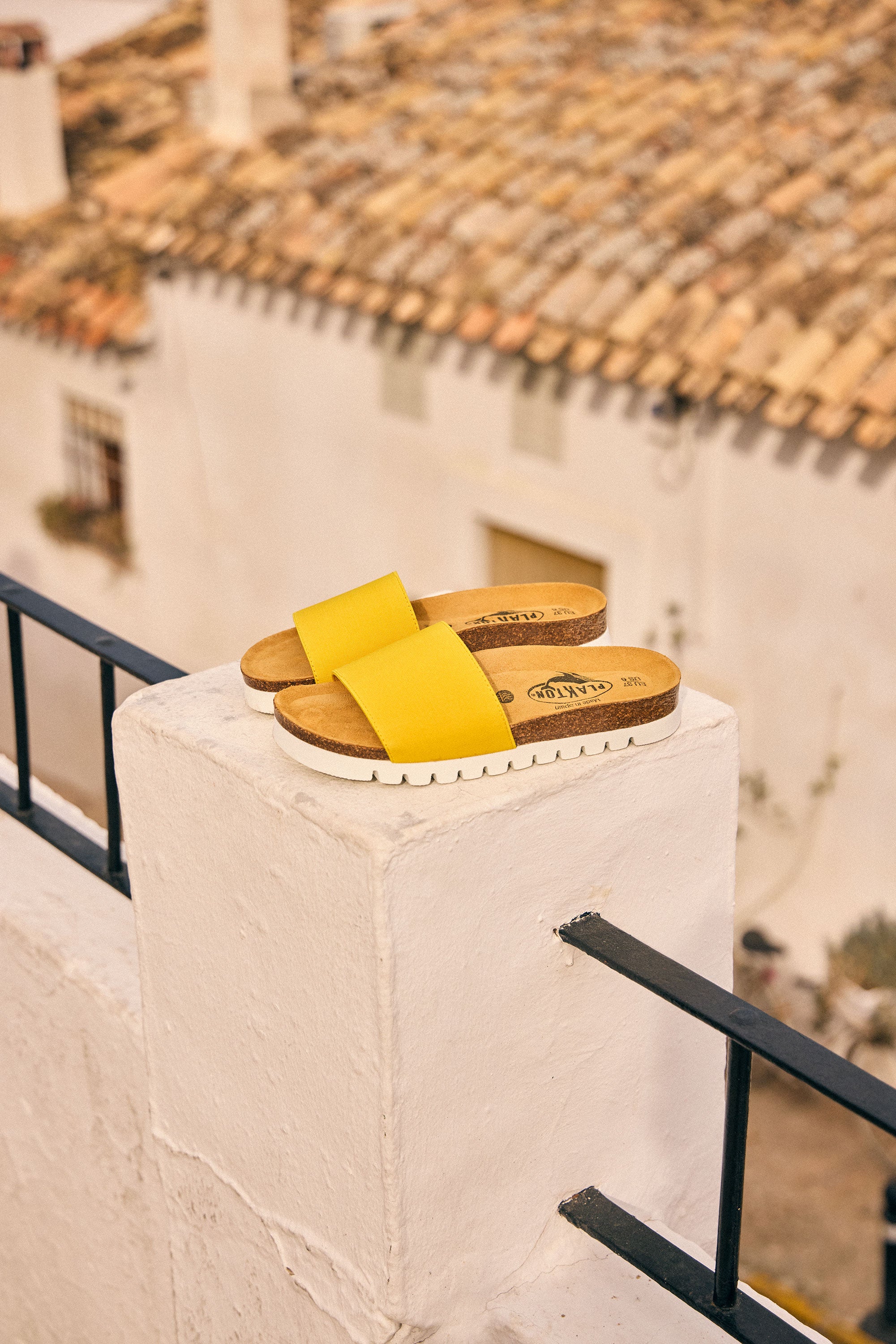 Plakton Barcadi yellow flat sandals with cork footbed and white sole placed on a white balcony wall overlooking a charming Spanish village, highlighting comfortable women’s summer sandals handmade in Spain.