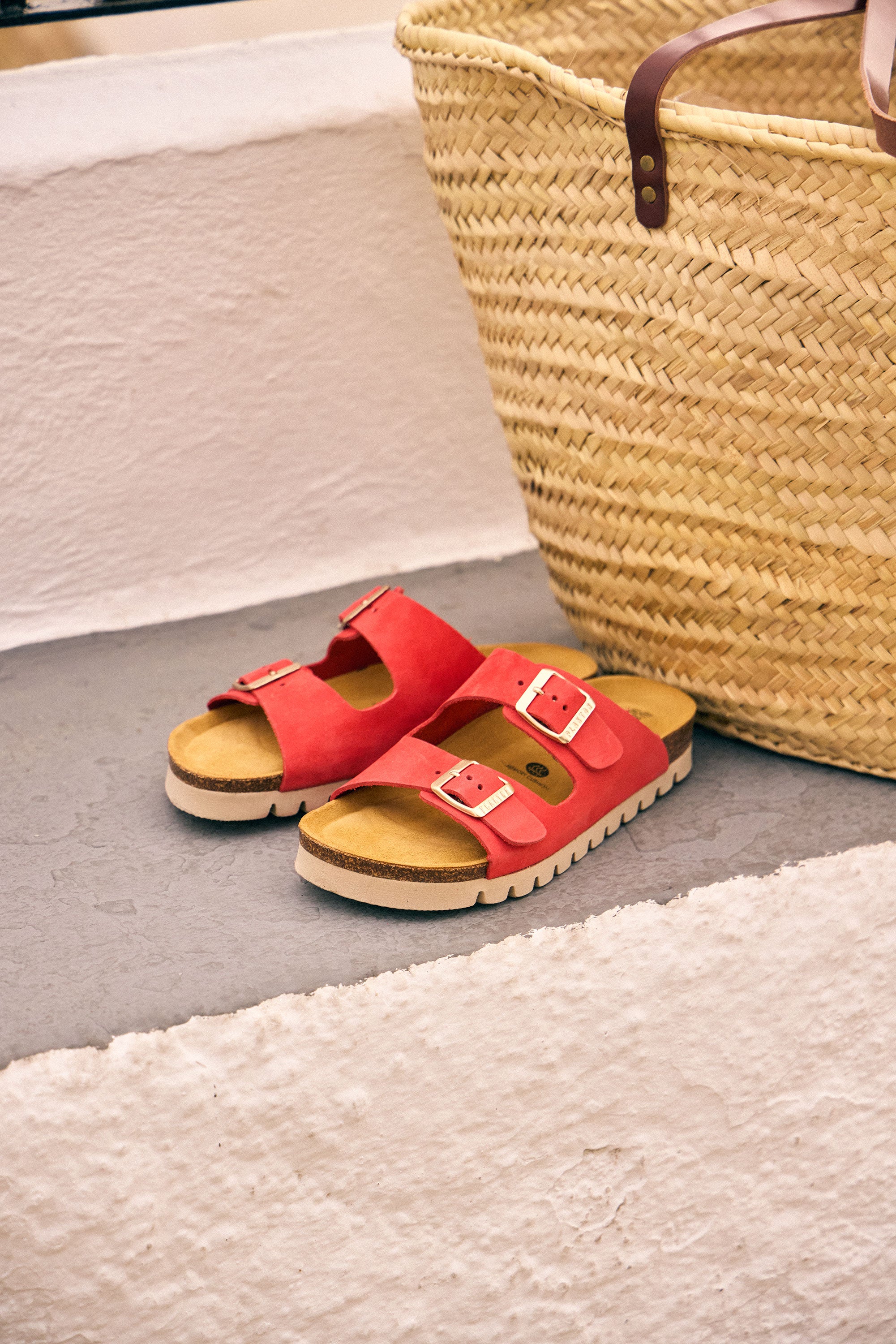 Plakton Beta coral two-strap cork footbed sandals with adjustable buckles placed beside a woven straw basket on white stone steps, showcasing comfortable handmade summer sandals from Spain.