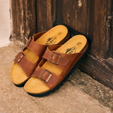 Plakton Beta two-strap brown sandals with adjustable buckles and cork footbed resting against a rustic wooden door in a Spanish village, showcasing handmade comfort sandals with a 15 mm sole.