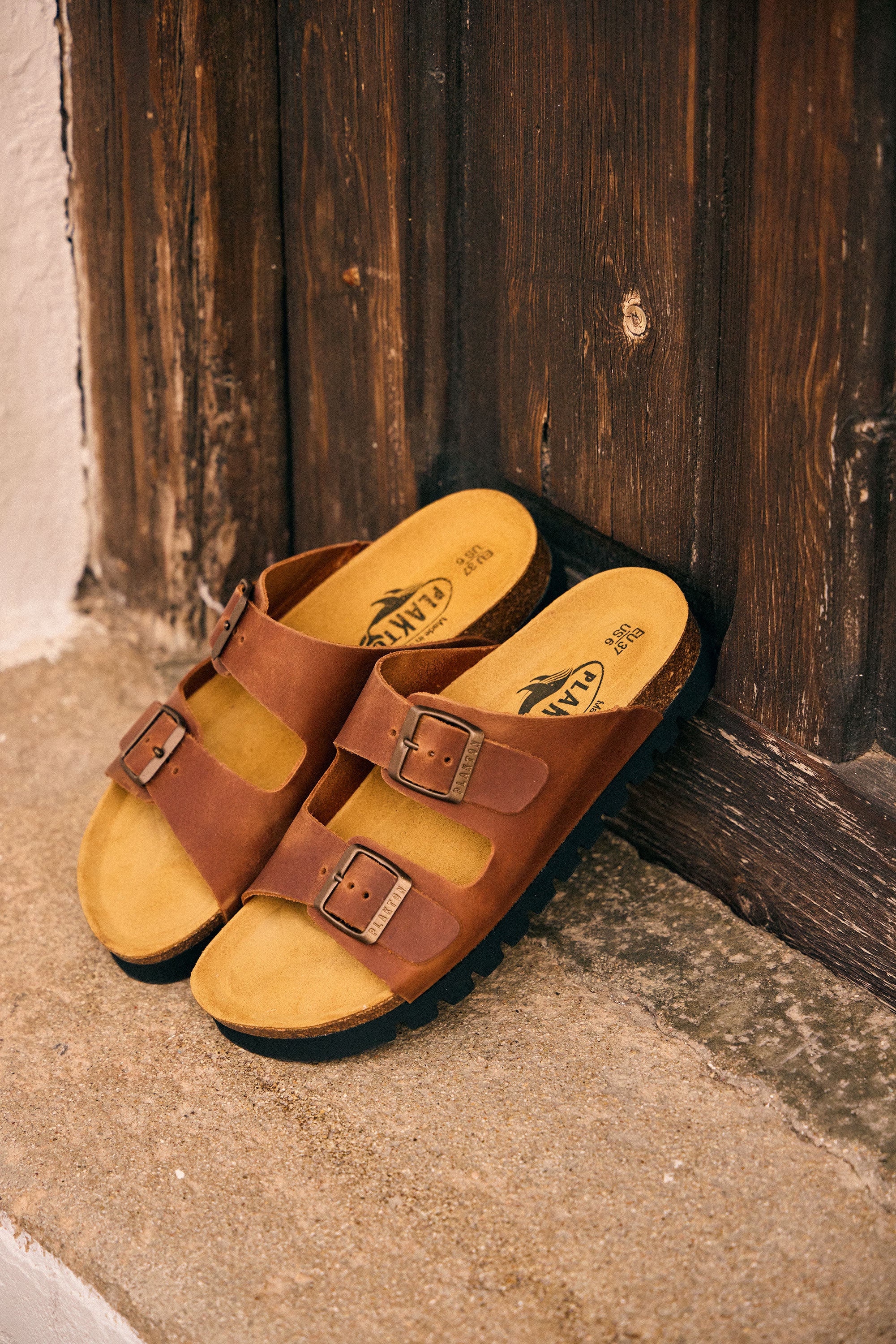 Plakton Beta two-strap brown sandals with adjustable buckles and cork footbed resting against a rustic wooden door in a Spanish village, showcasing handmade comfort sandals with a 15 mm sole.