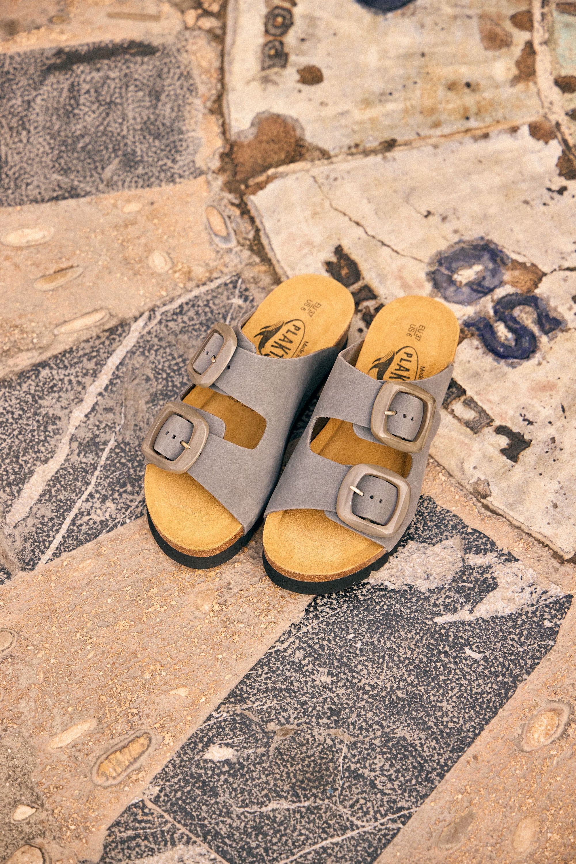 Plakton Calla two-strap sandals in taupe with adjustable buckles and cork footbed placed on patterned stone pavement, highlighting comfortable handmade summer sandals from Spain.
