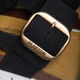 An extreme close-up focusing on the fine, matte texture of the Black nubuck leather. A sturdy metallic buckle embossed with "PLAKTON" anchors the strap, while the soft-focus background reveals the porous, breathable texture of the natural Spanish cork, highlighting the union of high-quality hardware and eco-conscious craftsmanship.