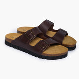 A pair of Barna Sandals in Coffee Dakota Leather, showing the dual adjustable straps with antique-finish metallic buckles. The deep, cupped heel of the natural cork footbed is lined in soft tan suede, showcasing a design that balances timeless Mediterranean style with the wisdom of anatomical support.