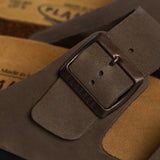 An extreme close-up of the Barna’s upper strap. The focus is on the matte, velvety grain of the khaki nubuck and a sturdy bronze buckle embossed with "PLAKTON." In the blurred background, the porous texture of the Spanish cork and the plush suede lining reinforce the premium, artisan quality of these Spanish-made sandals.