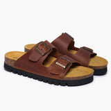 A pair of Beta sandals in Brown Oil Leather, angled to reveal the deep heel cups and the smooth transition of the handcrafted cork base. The dual adjustable straps feature antique-finish metallic buckles, resting atop a plush tan suede lining that highlights the artisan "Nature’s Hug" philosophy.