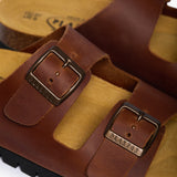 An extreme close-up of the Beta strap, focusing on the heavy-grain and pull-up effect of the Brown Oil Leather. A sturdy metallic buckle embossed with "PLAKTON" is the centerpiece, while the soft-focus background highlights the porous, sustainable cork, emphasizing the premium "Handmade in Spain" heritage.