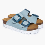 A pair of Calla sandals angled to highlight the deep, anatomical heel cups carved from renewable cork oak. The dual adjustable Light Blue straps feature brushed metallic buckles and rest atop a plush tan suede lining, embodying the "Nature’s Hug" philosophy—where handcrafted detail meets the raw purity of the earth.